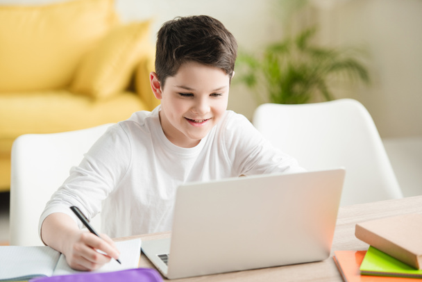 cheerful boy using laptop and writing in copy book while doing schoolwork at home - Foto, Bild