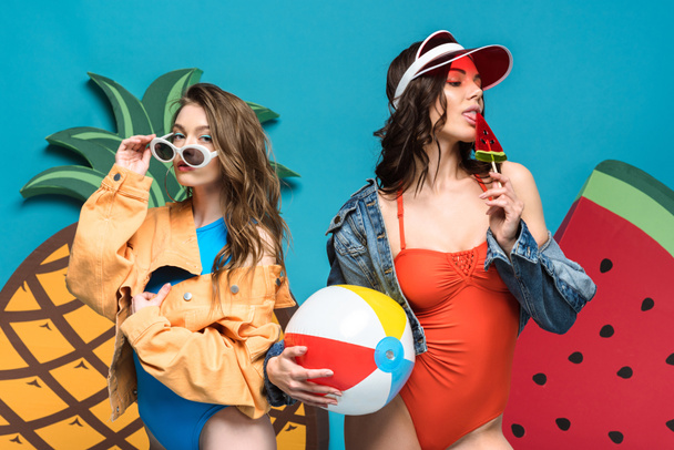 dvě dívky s plážovým míčkem a lízátka blízko dekorativního melounu a ananasu izolovaně na modrém - Fotografie, Obrázek