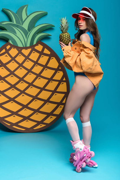 full length view of sexy girl in roller skates holding pineapple on blue - Fotoğraf, Görsel