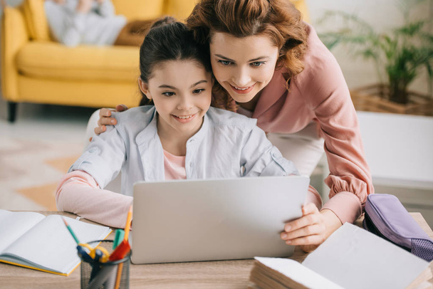 happy mother embracing smiling daughter doing schoolwork at home - Zdjęcie, obraz