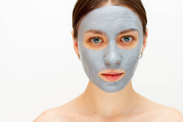 Portrait of young lady is looking at camera with joy while touching her face with clay mask while using procedures. white background. Beauty concept - Photo, Image