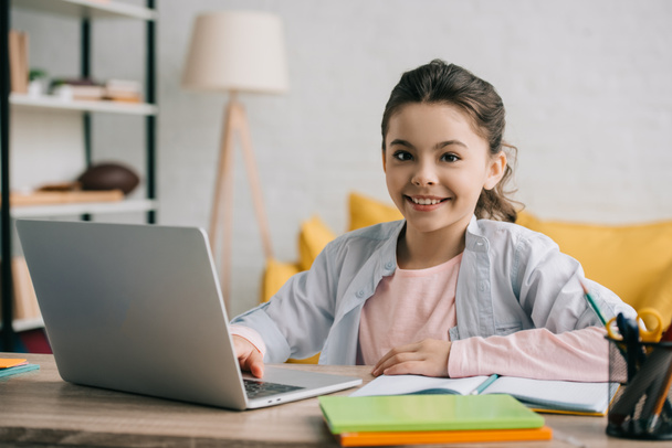 счастливый ребенок смотрит на камеру, сидя за письменным столом и пользуясь ноутбуком дома
 - Фото, изображение