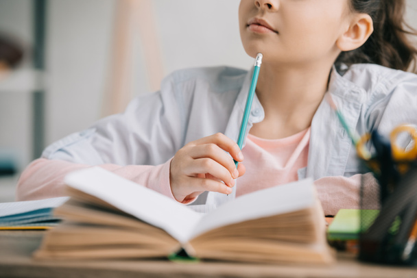 vista parcial del escolar sosteniendo el lápiz mientras está sentado en el escritorio cerca del libro y haciendo la tarea
 - Foto, Imagen