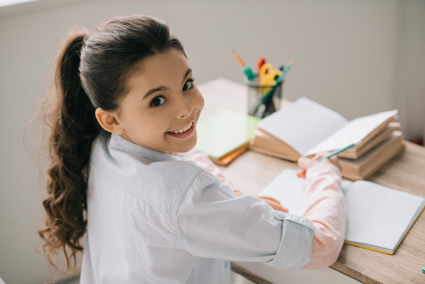 fröhliches Schulkind, das am Schreibtisch in die Kamera schaut und Hausaufgaben macht - Foto, Bild
