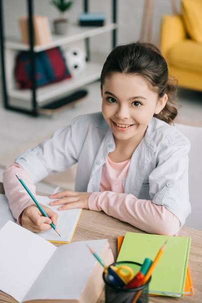 cheerful schoolkid writing in notebook and smiling at camera while doing homework - Foto, Bild