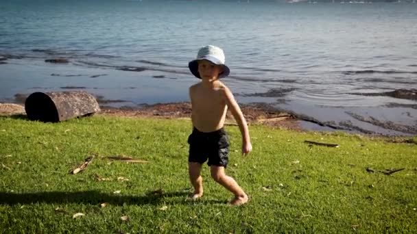 Młody chłopiec w szortach i kapeluszu wiadro na trawiastym brzegu jeziora na wysokości lata pokazując swoją wersję jak zrobić "Nić dentystyczna" - Materiał filmowy, wideo