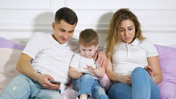 Семья с ребенком смотрит в свои мобильные телефоны, лежащие на кровати. Зависимость от гаджетов
. - Фото, изображение