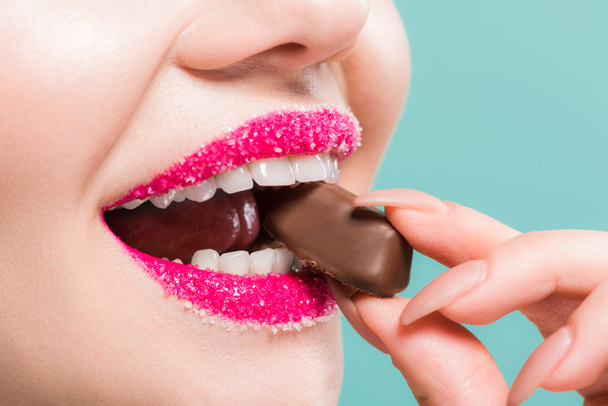青で隔離されたチョコレートキャンディを食べて唇に砂糖を持つ女性のトリミングビュー   - 写真・画像