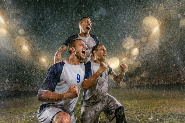 Jugadores de fútbol en el estadio profesional de fútbol nocturno de lluvia. Tres jugadores sucios bajo la lluvia gotas emocionalmente regocija la victoria. Los hombres se arrodillan y gritan
 - Foto, imagen