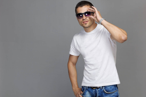 Beautiful close-up of a guy in a white cotton t-shirt and sunglasses on a gray background. Wrist watch isolated background - Zdjęcie, obraz