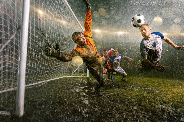 サッカー選手は、プロの夜の雨のスタジアムでアクションプレイを行います。雨の中の汚れた選手は、頭でゴールを決めます。ボールをキャッチしようとする飛行中のゴールキーパー。雨で濡れたスタジアムの草 - 写真・画像