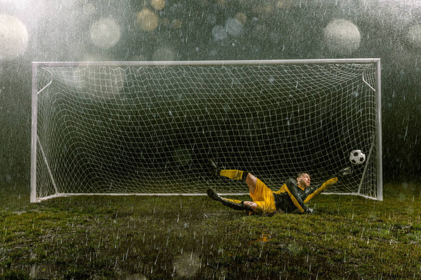 Ο βρώμικος τερματοφύλακας στην πτήση πιάνει την μπάλα. Επαγγελματικό νυχτερινό στάδιο βροχής με στόχο το ποδόσφαιρο. Γρασίδι στο στάδιο υγρό από τη βροχή - Φωτογραφία, εικόνα