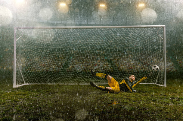 Dirty goalkeeper in flight catch the ball. Professional night rain stadium with football goal. Grass in the stadium wet from the rain - Photo, Image