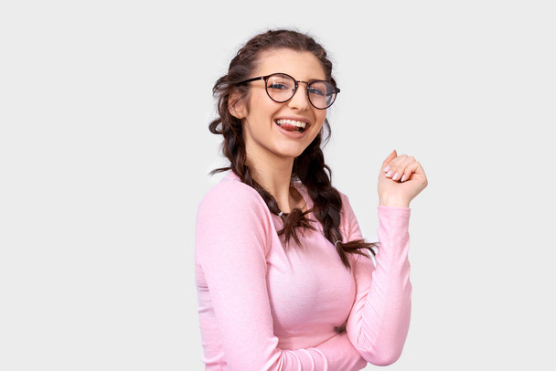 Pozytywna cute młoda kobieta uśmiecha się szeroko, nosi różowy casual bluzka i okrągłe okulary przezroczyste, izolowane na białym tle. Szczęśliwa młoda kobieta z warkocze fryzurę na szarym ścianie Studio. - Zdjęcie, obraz