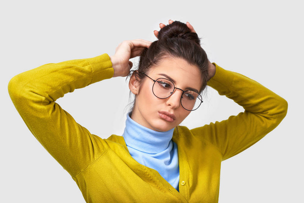 Shot of cute young woman with healthy clean skin, wearing yellow and blue sweater, making a knot hairstyle, with serious expression. Caucasian female with hair gathered in bunch posing indoors. - Photo, Image