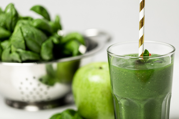 selective focus of tasty green smoothie in glass with straw near spinach leaves and apple on white  - Photo, Image