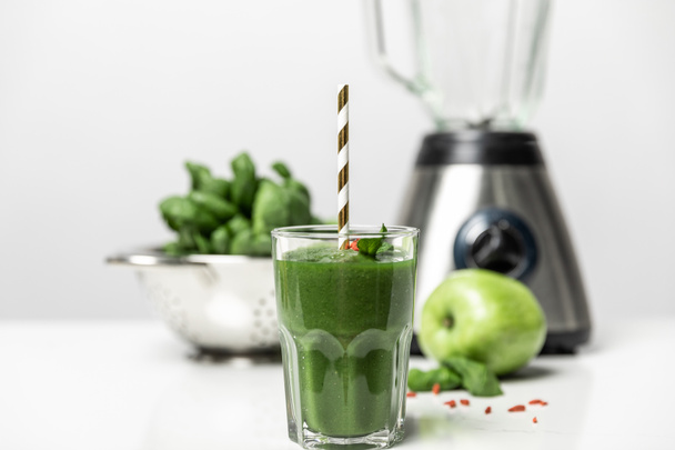 selective focus of tasty smoothie in glass with straw near fresh spinach leaves, apple and blender on white  - Photo, Image