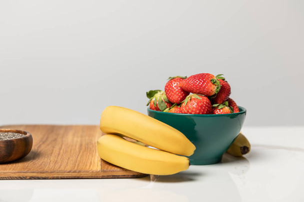 sweet and tasty strawberries in bowl near yellow bananas and cutting board on grey  - Photo, image