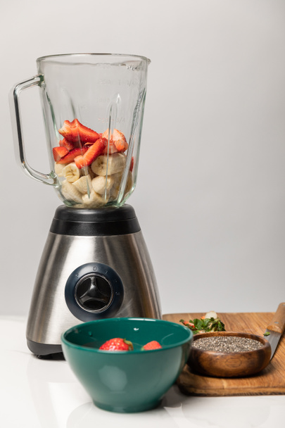 sweet strawberries and ripe bananas in blender near bowls and cutting board on grey  - Photo, Image