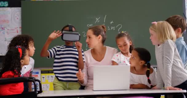 Front view of African american schoolboy using virtual reality headset with teacher and classmates in the classroom.  Female teacher using laptop  - Materiaali, video