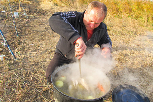 Мужчина готовит суп шурпа в большом чугунном котле на открытом воздухе. Летняя еда для походов, рыбалки
 - Фото, изображение