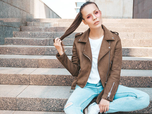 Beautiful brunette model dressed in summer hipster jacket and jeans clothes. Trendy girl sitting on steps in the street background. Funny and positive woman in round sunglasses. Playing with hair - Photo, image