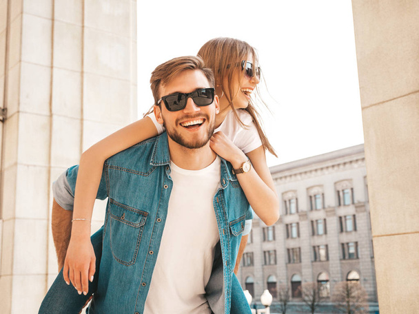 Smiling beautiful girl and her handsome boyfriend in casual summer clothes. Man carrying his girlfriend on the back and she raising her hands.Happy cheerful family having fun on the street background  - Fotó, kép