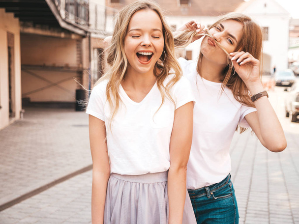 Moda yaz beyaz t-shirt giysiler iki genç güzel sarışın gülümseyen hipster kız Portresi. Seksi kaygısız kadınlar sokak arka planda poz. Eğlenen pozitif modeller - Fotoğraf, Görsel
