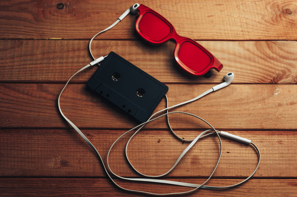 Kırmızı gözlük, klasik manyetik kaset ve kahverengi ahşap masa arka plan üzerinde beyaz kulaklıklar. - Fotoğraf, Görsel
