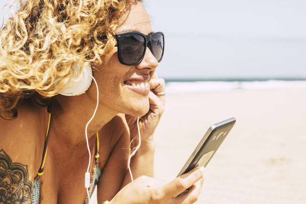 hübsche Mädchen mit lockigen Haaren und kurzen Jeans genießen am Strand während Musik hören  - Foto, Bild