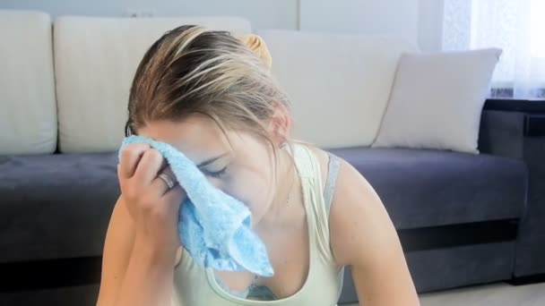 Slow motion footage of exhausted young woman sitting on fitness mat and wiping sweat with towel from her forehead after exercising - Imágenes, Vídeo