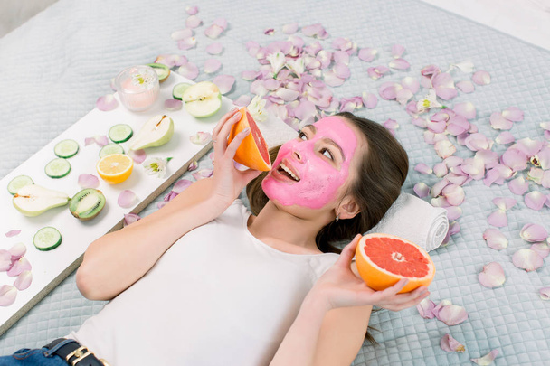 portrait of pretty young woman with pink clay mask on her face holding slices of grapefruit, facial care. - Fotó, kép