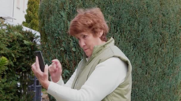 Seniorin fotografiert mit Handy im Garten, spricht und lächelt draußen, Tag. Lachende Großmutter mit Smartphone im Park. Eine ältere Dame macht ein Selfie mit dem Handy. Aktiver Lebensstil im Alter, draußen. Ältere Person fotografieren auf Gadget - Filmmaterial, Video