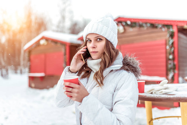 Beautiful girl in the winter in the city, calls on phone, in a white hat and jacket. In his hand holds a cup of coffee tea. Free space for text. - Foto, Imagen