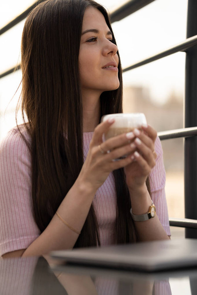 Το κορίτσι απολαμβάνει καφέ. Ένα φλιτζάνι καφέ σε γυναικεία χέρια. Στο τραπέζι είναι ένας κλειστός φορητός υπολογιστής - Φωτογραφία, εικόνα
