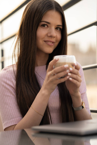 Kız kahvenin tadını çıkarıyor. Kadın ellerinde bir fincan kahve. Masada kapalı bir dizüstü bilgisayar var. - Fotoğraf, Görsel