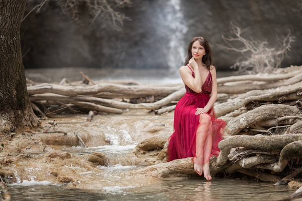 Young beautiful woman is wearing fashion red dress sitting in forest near the waterfall - 写真・画像