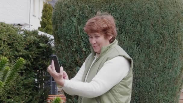 Mujer mayor sonriente recibiendo selfie por teléfono móvil en el jardín, día. Abuela usando smartphone en el parque. Anciana sacando fotos por celular al aire libre. Estilo de vida activo de anciano, afuera. Persona mayor tomar fotos en gadget
 - Metraje, vídeo