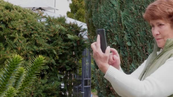 Gülümseyen yaşlı kadın bahçede fotoğraf çekiyor ve cep telefonuyla selfie çekiyor. Büyükanne parkta akıllı telefon kullanıyor. Yaşlı bayan açık havada cep telefonuyla fotoğraf çekiyor. Yaşlıların aktif yaşam tarzı, dışarıda. Yaşlı kişi gadget'ta resim çekmek - Video, Çekim