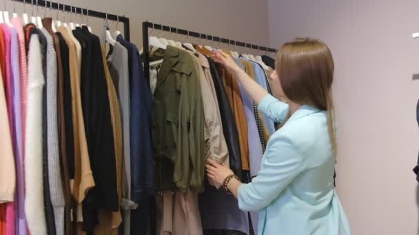 menina loira atraente escolhe roupas olhando através de amostras penduradas em racks na loja moderna câmera lenta
 - Filmagem, Vídeo
