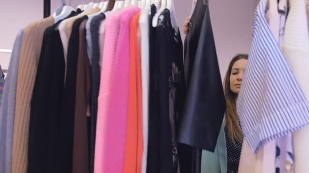 dość długowłosy dziewczyna szuka sukienka zamknąć widok przez ubrania wiszące na stojakach w nowoczesnym sklepie powolny ruch - Materiał filmowy, wideo