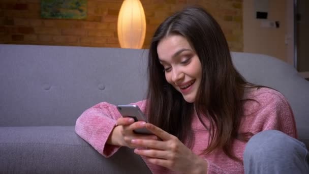 Closeup shoot of young happy brunette caucasian female using the phone and smiling sitting on the floor in a cozy apartment indoors - Footage, Video