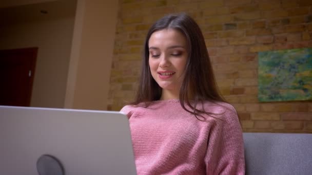 Closeup shoot of young pretty brunette caucasian female student using the laptop sitting on the couch in a cozy apartment indoors - Filmmaterial, Video