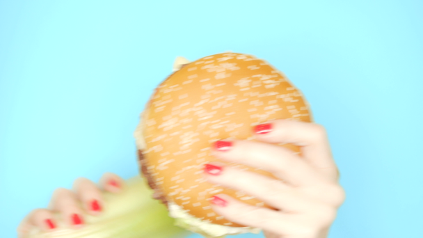 Concept of healthy and unhealthy food. celery stalks against hamburgers on a bright blue background. female hands with red nail polish hold burger and celery - Filmagem, Vídeo