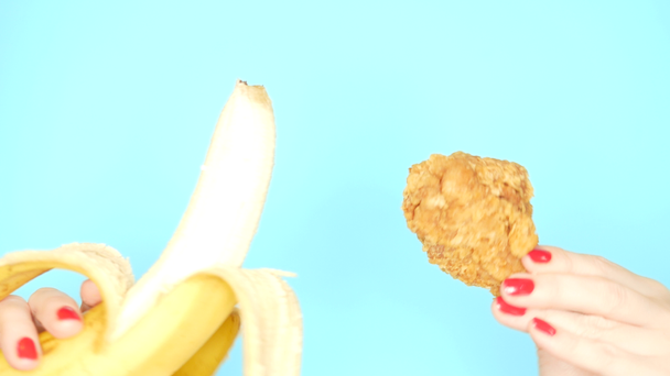 Koncept zdravého a nezdravého jídla. banán proti smaženým obaleným kuřecím nožům na jasně modrém pozadí. ženské ruce s červeným leštěkem na nehty drží banán a Nuggets - Záběry, video