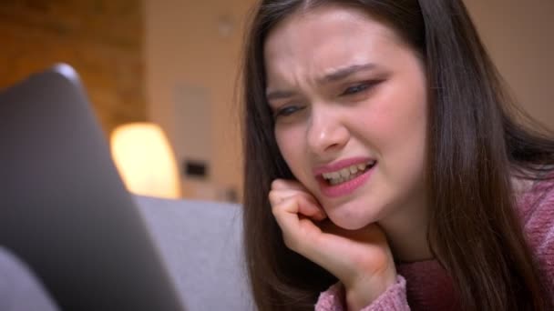 Closeup shoot of young pretty brunette caucasian female teenager having a video call on the laptop and reacting with disgust on the couch in a cozy apartment indoors - Materiaali, video