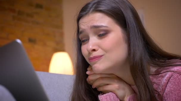 Closeup shoot of young pretty brunette caucasian female teenager having a video call on the laptop and being amused on the couch in a cozy apartment indoors - Filmagem, Vídeo