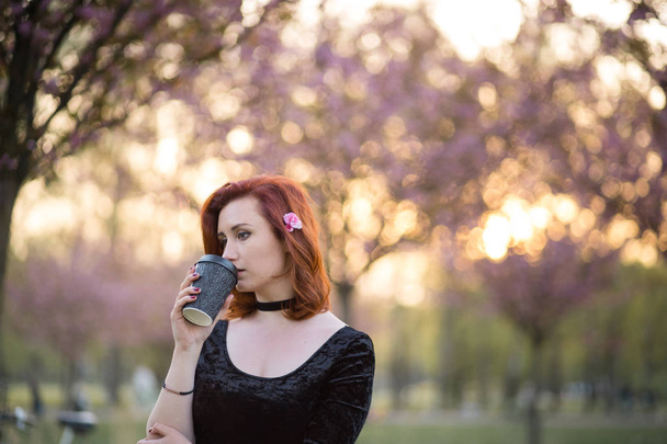 Πίνοντας καφέ από χαρτί κούπα Κύπελλο-ευτυχής νεαρή χορεύτρια ταξίδι που απολαμβάνουν ελεύθερο χρόνο σε ένα πάρκο ανθοφορία κερασιάς-Καυκάσιος λευκό κοκκινομάλλα κορίτσι - Φωτογραφία, εικόνα