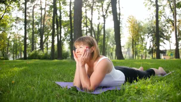 4k Zeitlupenaufnahmen einer lächelnden Frau mittleren Alters, die an einem sonnigen Sommertag im öffentlichen Park Yoga praktiziert. Konzept der körperlichen und geistigen Gesundheit - Filmmaterial, Video