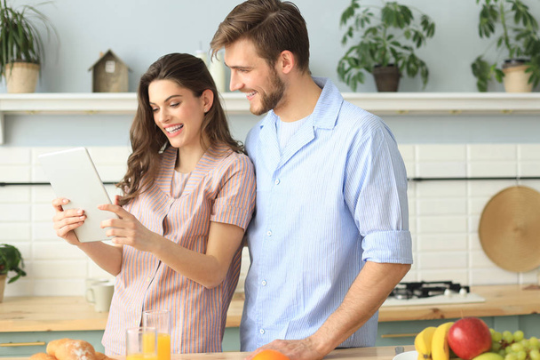 Happy young couple in pajamas watching online content in a tablet and smiling while cooking in kitchen at home. - Valokuva, kuva